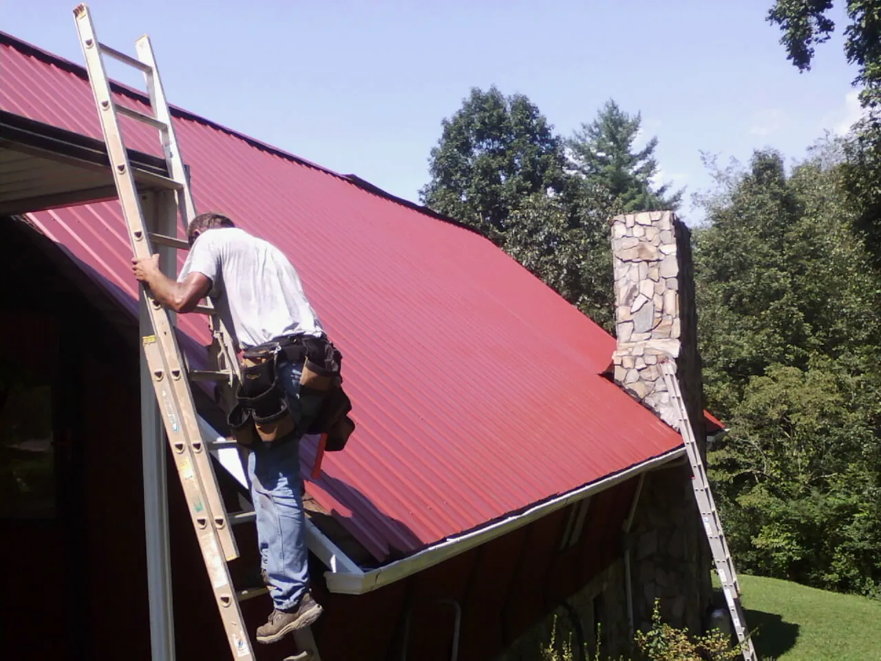 Roofer on a ladder beside a red metal roof and stone chimney — Soffit & Fascia Repair by Roofing Brothers in Hamptonville